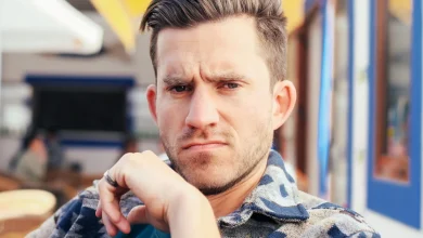 Man with short brown hair and stubble frowning skeptically at the camera with his hand at his chin, seated at an outdoor cafe.
