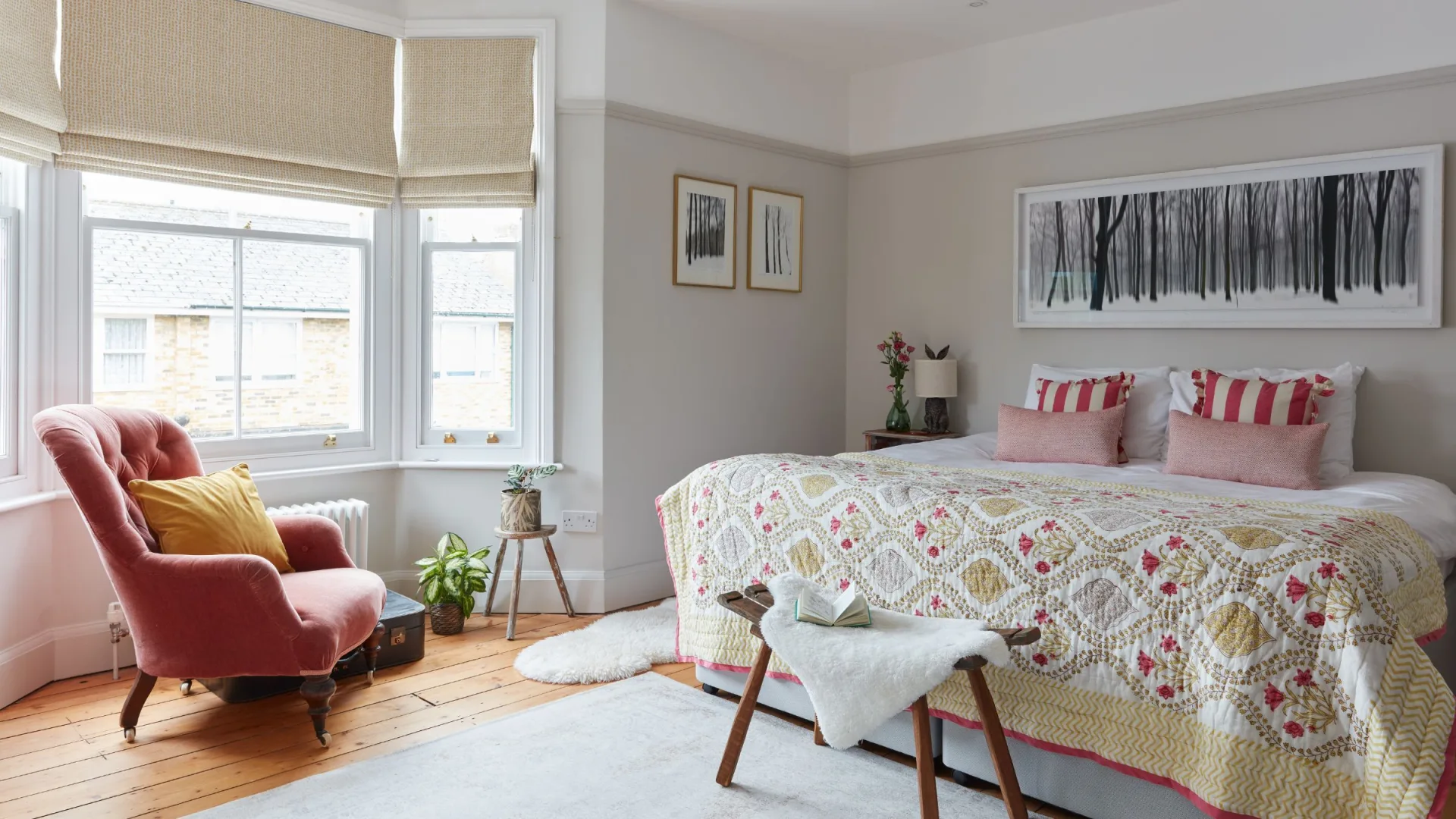 neutral painted period bedroom with bay window and a pink accent chair in front of it, and a large bed on the right hand…