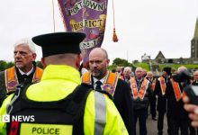 A police officer with his back turned to the camera. There is a group of orange men marching along the road next to a…