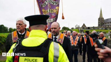 A police officer with his back turned to the camera. There is a group of orange men marching along the road next to a…