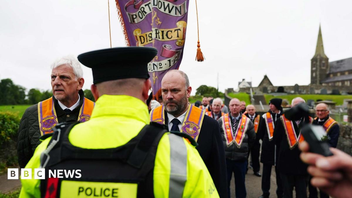 A police officer with his back turned to the camera. There is a group of orange men marching along the road next to a…
