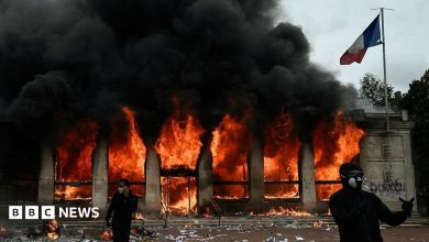 Two demonstrators, dressed all in black and in face coverings, stand outside the Lyon tourist office which is on fire behind them. A French…