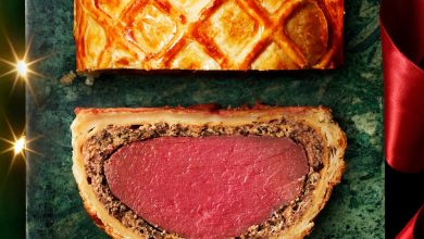 A slice of beef Wellington showing pink beef wrapped in mushroom filling and golden pastry, beside the remaining lattice-topped Wellington.