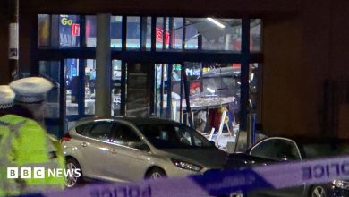 A car can be scene in the background inside a store with damage to the shop windows. A silver car is parked outside the…