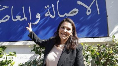 El Mansouri, wearing a black blazer over a white shirt, stands in front of her party's slogan, written in Arabic in white lettering against…