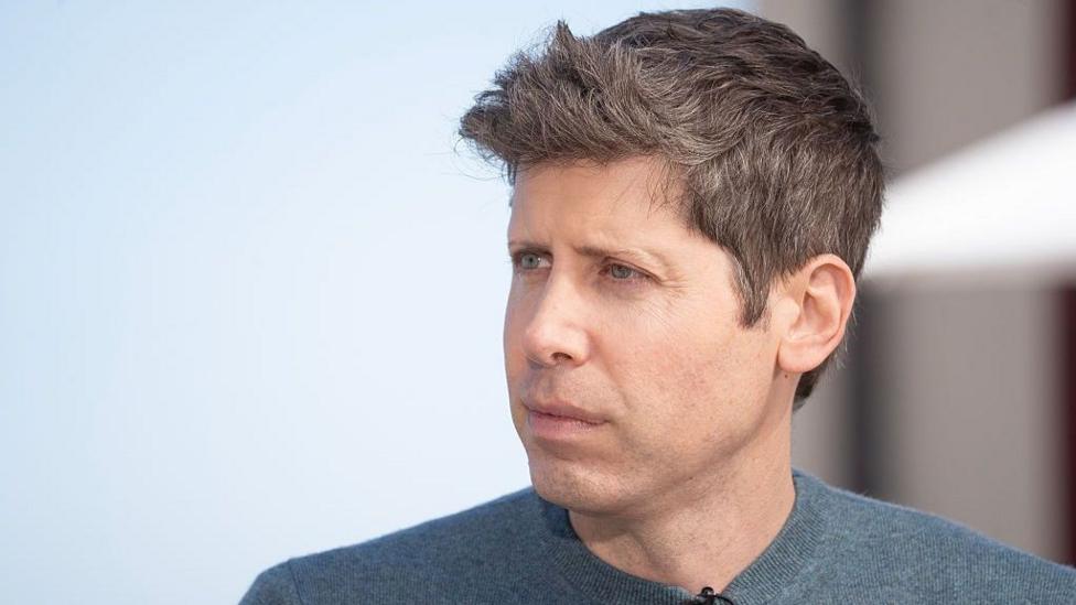 Sam is wearing a grey-blue sweater and looks serious in a close up image.