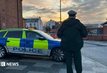 A police officer stands with his back to the camera. He is in uniform and standing beside a police car. Police tape can be…