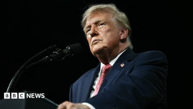 President Trump stands against a black background behing a black podium/ He is wearing a white shirt, blue suit coat with an American flag…