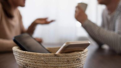 Two smartphones sit in a woven basket on a table as people talk in the background, signaling a screen-free day.