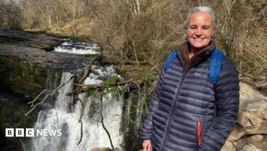 Eleanor stands by a waterfall. She has white hair, which is tied back, and wears a navy coat. She is smiling at the camera.